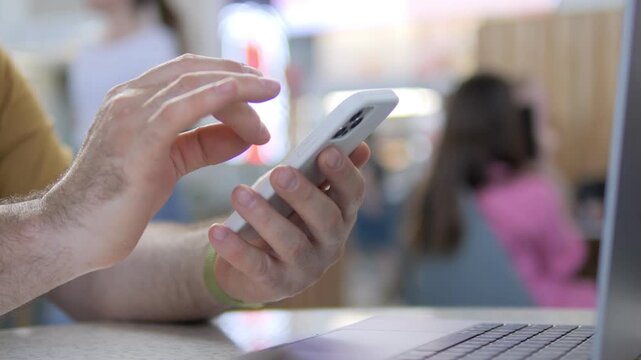 Person using smartphone while sitting at table. Fingers scroll through phone screen actively. Laptop beside phone, suggesting multitasking. Background shows blurred people, implying public setting