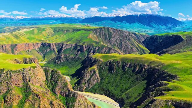 Scenic aerial view of Kuoksu Grand Canyon featuring lush green grasslands, steep cliffs, a winding river, and snow-capped Tien Shan mountains in Xinjiang, China.