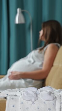 Expectant mother sitting on a couch caressing her pregnant belly while anticipating childbirth, with a focus on newborn clothes and booties prepared for the baby's arrival