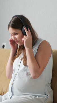 Content pregnant woman with a big belly sitting on a couch, putting on wireless headphones and happily listening to music while gently moving to the beat, enjoying a moment of relaxation