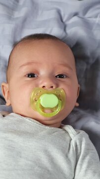Charming newborn baby with a green pacifier lying on a bed and looking at the camera. Sweet and innocent infant with big dark eyes smiling, capturing a peaceful moment of early childhood