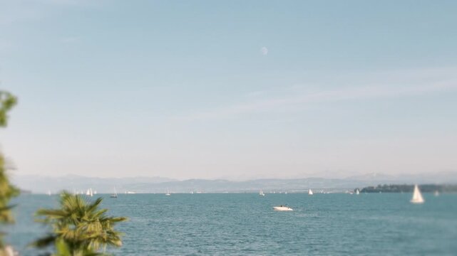 Slow motion widescreen view of Lake Constance with sailboats, a hazy mountain horizon, palm tree and daytime moon, real footage for website and desktop backgrounds