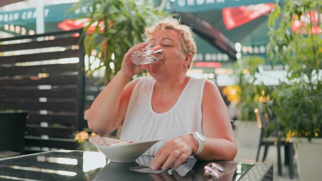 Senior overweight blonde woman drinking fresh water from glass at outdoor restaurant.