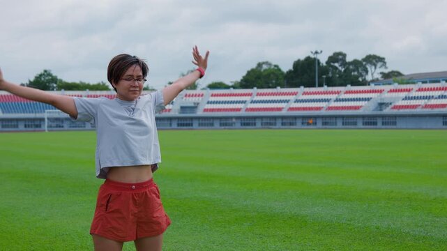Authentic shot of an active Asian woman stretching her arms on a green soccer field. This empty stadium with colorful seats provides excellent copy space for your sports backgrounds.
