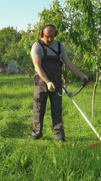 Gardener in gray overalls uses a string trimmer to cut grass in a lush green garden, surrounded by trees and a clear blue sky, showcasing the progression of lawn maintenance tasks