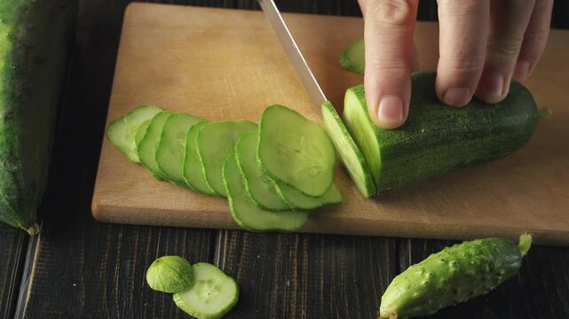 Close-up of hands slicing fresh cucumber into thin rounds on a wooden cutting board, showcasing the continuous action of cutting with a sharp knife and the vibrant green color of the vegetable
