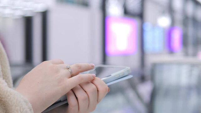 Person holds tablet, taps screen. Background shows blurred airport or transit area. Pink and blue lights glow behind. Hands appear steady, focused on device