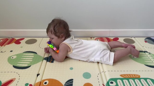 Infant lies on stomach playing with plastic rattle toy on colorful soft play mat indoors. Early child development focus highlights motor skills growth sensory discovery and playful learning at home.