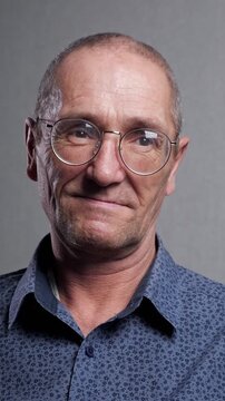 Close up portrait of a pensive senior caucasian man wearing round eyeglasses and a blue shirt, looking directly at the camera with a subtle, gentle smile against a gray studio background
