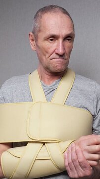 Worried mature man with a fractured arm wearing a medical sling and gently touching his injured wrist, experiencing severe pain and discomfort during his rehabilitation process