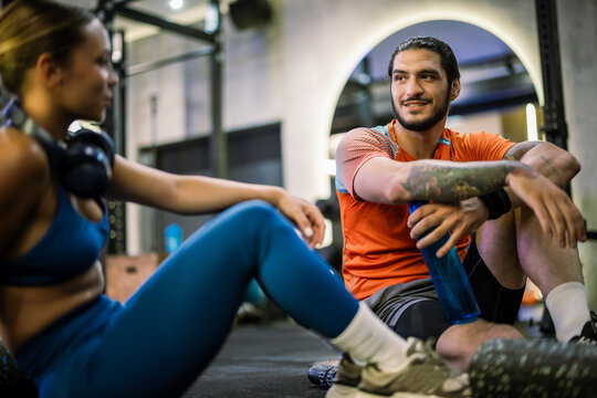 Athletic couple resting on gym floor with protein shakes and headphones around neck