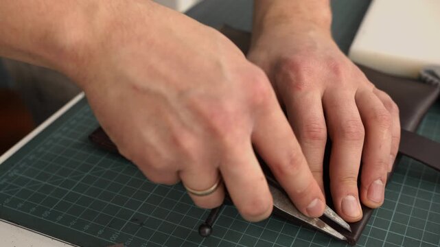 Leatherworker using a compass tool for marking a seam line on a handmade brown leather wallet in a workshop