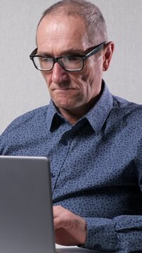 Mature caucasian businessman wearing glasses diligently working on his laptop, demonstrating concentration and focus while typing on the keyboard against a simple gray background