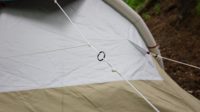 A close-up shows a white guy line cord threaded through a metal ring and secured with knots on a camping tent. Concept of outdoor equipment assembly, camping preparation, and secure shelter.