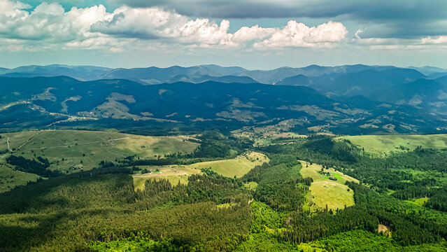 Aerial view of rolling mountains and sunlit valleys with forests and open meadows under dramatic clouds and blue sky showing the vast peaceful beauty of a remote highland landscape