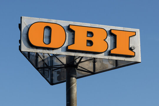 Weithin sichtbares Firmenschild des Baumarkts OBI auf einem Pylonmast vor blauem Himmel. Das orange-silberne Logo ist das markante Erkennungszeichen der Baumarktkette.