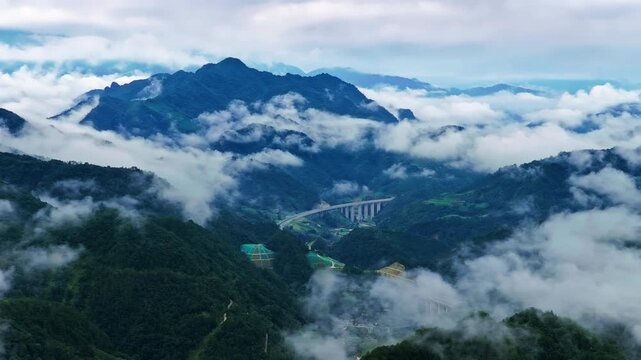 Highway bridge stretching through foggy mountain valley surrounded by lush green peaks and low clouds.