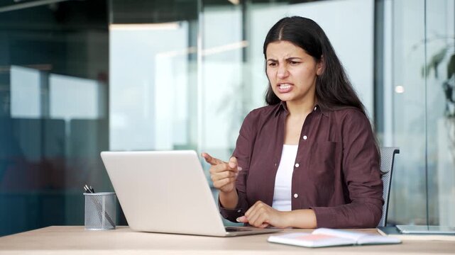 Angry businesswoman arguing during online meeting on laptop while sitting at desk in office. Disappointed female employee quarrel, dealing with work problems, reacts emotionally on video call