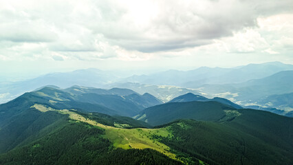 Fototapeta premium Sunlight pours across a sawtooth ridge then slides into shadowed hollows. The view flows over dark pines toward remote towns and river valleys framed by sweeping hills and silver skies
