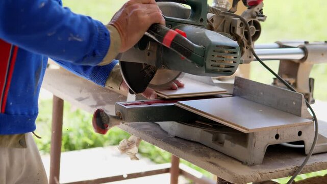 Close-up side view of carpenter in blue jacket steadying flat wood boards under bench mounted miter saw, with hand on material and other hand on tool, in bright outdoor workshop