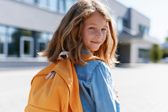 Young girl smiles softly, backpack slung over shoulder. She wears denim jacket, bright orange satchel. School building looms in background, windows gleaming. Sunlight bathes scene, casting soft