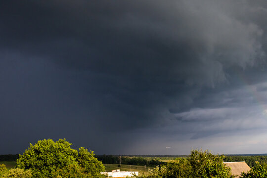 An advancing storm edge swallows the landscape while brighter skies retreat to the right green fields and trees still catch sun as the dark mass signals an imminent burst of rain