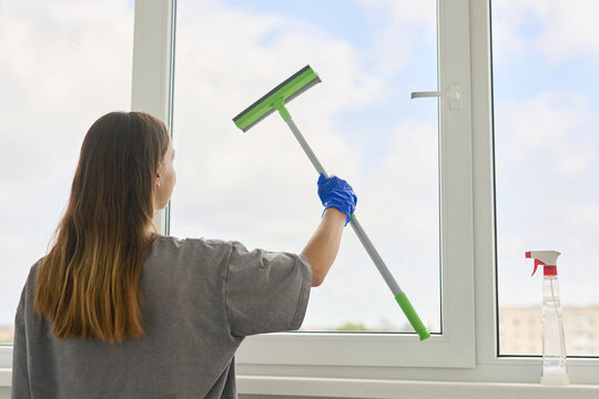 Cleaning lady wearing blue gloves washing a window glass with a squeegee and detergent spray bottle