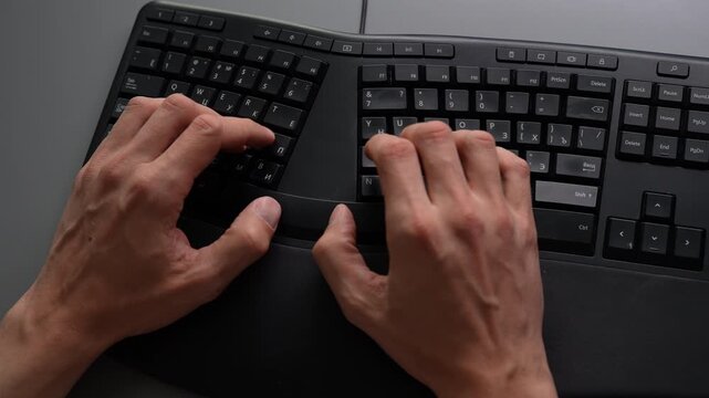 Top view of male hands typing on black ergonomic keyboard placed on gray office desk, illustrating concept of modern workflow, business communication, computer programming tasks, slow motion.