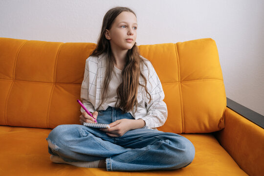 Pensive preteen girl with long hair sitting cross-legged on bright orange sofa, writing thoughts in diary. She looking away dreamily, holding pink pen. Casual, authentic lifestyle moment.