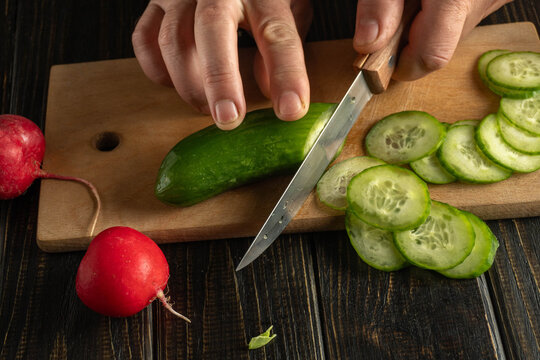 Hands are cutting cucumber on a wooden board while slices are piled next to a red radish. This scene takes place in a home kitchen during meal prep
