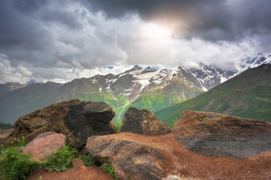 Mountains with snow peaks under dark storm clouds with sunlight. Dramatic horizontal landscape of high rocky view