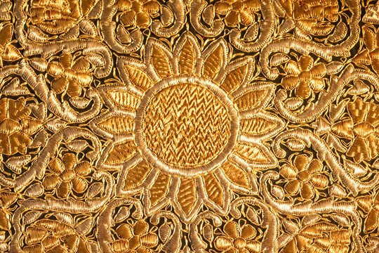Detailed golden embroidery showcases a floral design with a prominent sun motif at the center, set against a textured fabric background that enhances the intricate craftsmanship and artistry