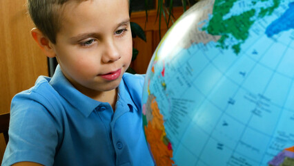 An eight-year-old boy sitting at a desk and exploring a globe while studying. Primary school education, geography learning, curiosity, homeschooling, classroom, and child development concept. © Sinelie