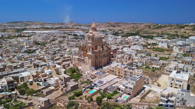 Xewkija, Gozo, Malta, with the Rotunda of St John Baptist Church rising above stone buildings during a village festa under blue sky, captured by drone.