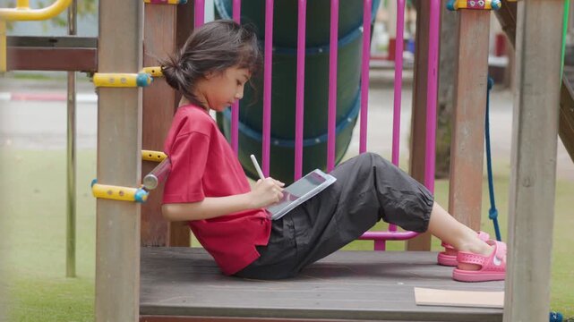 Asian kid enjoying a relaxing hobby, drawing digital art on tablet at outdoor school playground. Selective focus highlights creative elementary student spending personal leisure time with technology.