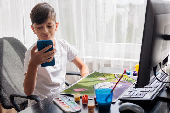 Young boy looking at a smartphone while painting at home. Concept of digital distraction, excessive screen time, reduced attention span, children's creativity, technology use, child development, balan