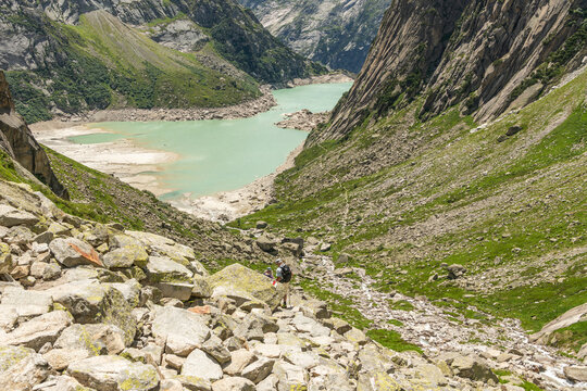 Guttannen, Switzerland. Hikers descending rocky alpine trail toward turquoise Gelmersee in the Bernese Alps