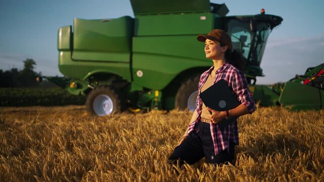 A farmer inspects crops in a field during late afternoon. A large green harvesting machine is parked nearby. The farmer holds a lap top computer and looks at the crops with focus.