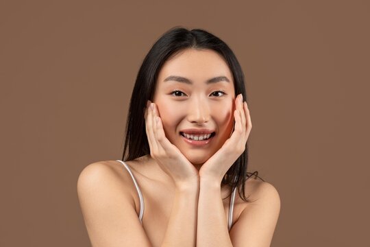 Happy Young Asian Woman Smiling And Showing Excitement With Hands On Face, Posing Against Brown Background In Studio. Reflecting Positive Emotions And Joyful Expression, Beauty Concept.