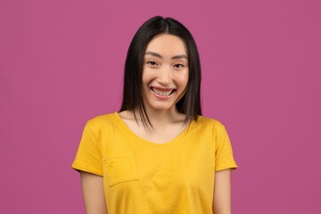 Fototapeta premium Happy Young Asian Woman With Braces Smiling At Camera, Wearing Yellow Shirt, Standing In Front Of Pink Background. Showing Joyful Emotion In A Fun, Colorful Setting. Lifestyle Concept.