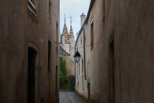 Narrow cobblestone alley in Beaune, France, during a rain shower, with historic buildings and the colorful tiled roof of a landmark Burgundy structure in the distance.