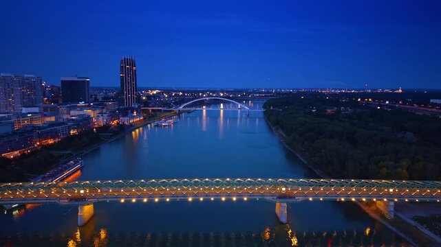 Illuminated Old Bridge across Danube River in Bratislava at night. Wide aerial perspective shows lit steel span, dark forest bank, and distant arch structure under blue sky.