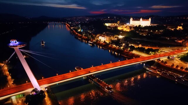 Aerial view of Most SNP bridge and Bratislava Castle at twilight. Illuminated bridge spans river while bright fortress stands on hill against dusk sky.