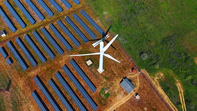 Top down aerial view of wind turbine and solar panels. White rotor blades cast a shadow across ground near rows of dark photovoltaic collectors.