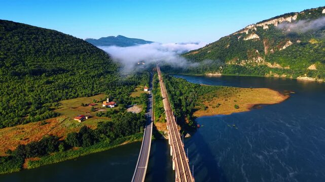 Aerial view of railway and road bridges across river valley. Morning fog drifts over dense green forest hills surrounding water and transport routes.