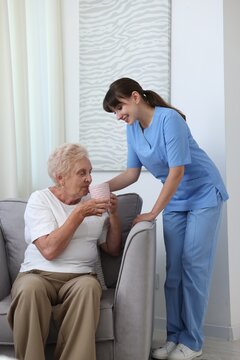Caregiver supporting elderly woman with cup of drink indoors