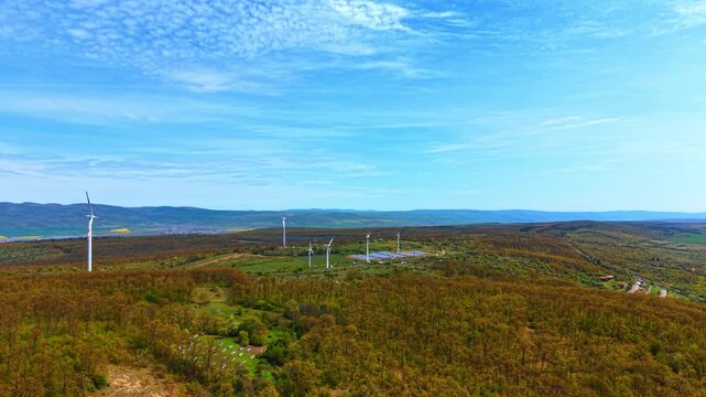 High aerial view of forest landscape with distant wind turbines. Dense green trees cover rolling hills while renewable energy structures stand in the far distance.