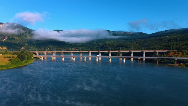 Parallel bridges spanning wide river in mountain valley. Low white clouds hang over green hills as morning mist drifts across dark blue water.