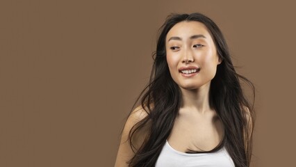 Fototapeta premium Happy Asian Woman Smiling With Long Hair, Looking To The Side In A Friendly Manner Against A Brown Background, Radiating Positive Vibes In A Studio Setting. Beauty Concept.