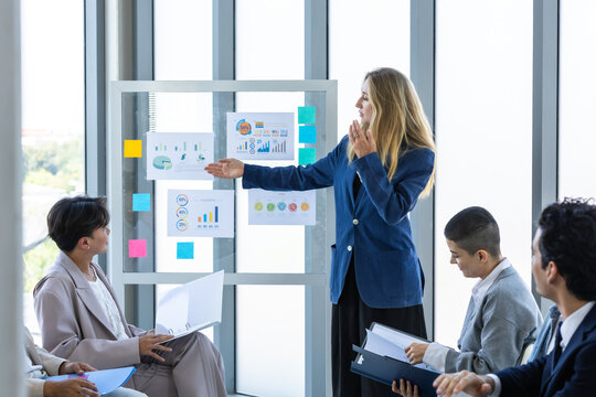 Confident businesswoman standing and presenting financial graphs on a glass board to her diverse team during a corporate strategy meeting in the meeting room at modern office.